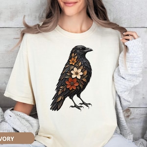 May include: An ivory short-sleeve t-shirt featuring a graphic illustration of a black crow adorned with orange, white, and brown floral patterns integrated into its feathers. The bird is depicted in profile facing right with detailed plumage.