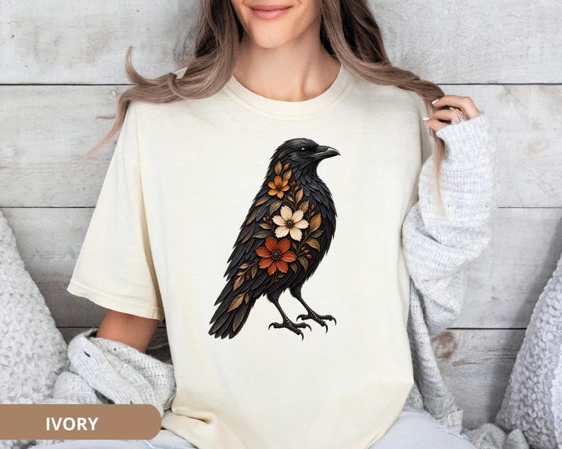 May include: An ivory short-sleeve t-shirt featuring a graphic illustration of a black crow adorned with orange, white, and brown floral patterns integrated into its feathers. The bird is depicted in profile facing right with detailed plumage.