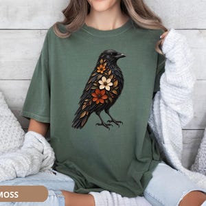 May include: A moss green short-sleeve t-shirt featuring a graphic print of a black crow with orange, white, and brown flowers integrated into its plumage. The shirt has a crew neckline and a relaxed fit, showcasing a detailed bird illustration.