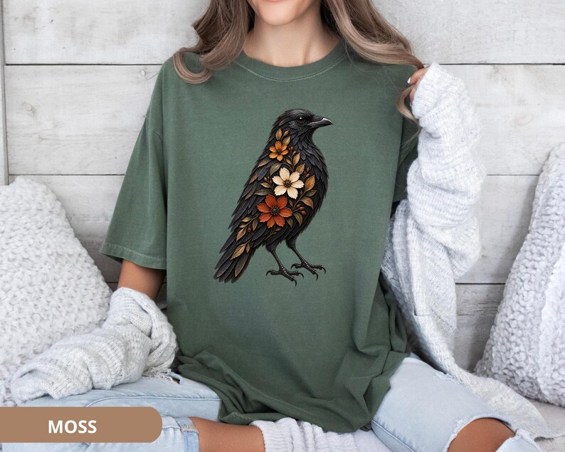 May include: A moss green short-sleeve t-shirt featuring a graphic print of a black crow with orange, white, and brown flowers integrated into its plumage. The shirt has a crew neckline and a relaxed fit, showcasing a detailed bird illustration.