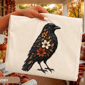 May include: An ivory-colored t-shirt featuring a graphic illustration of a black crow. The crow is decorated with a cluster of flowers in shades of orange, white, and brown on its chest and wing. The bird is depicted in profile facing right.