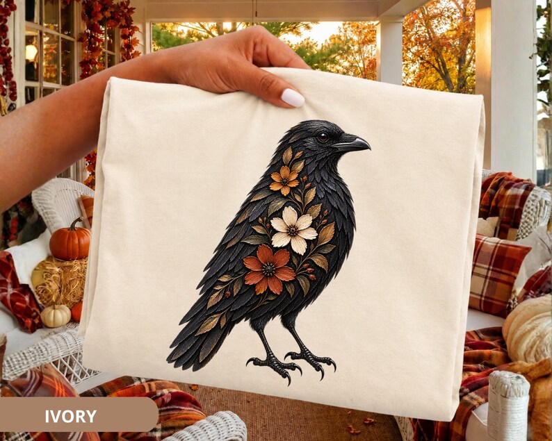 May include: An ivory-colored t-shirt featuring a graphic illustration of a black crow. The crow is decorated with a cluster of flowers in shades of orange, white, and brown on its chest and wing. The bird is depicted in profile facing right.