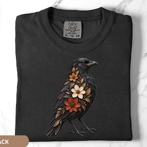 May include: A black t-shirt featuring a graphic of a crow with its chest filled with orange, white, and brown flowers and leaves. The shirt has a Comfort Colors brand label at the neckline with size XL-TG-EG printed on the interior fabric tag.