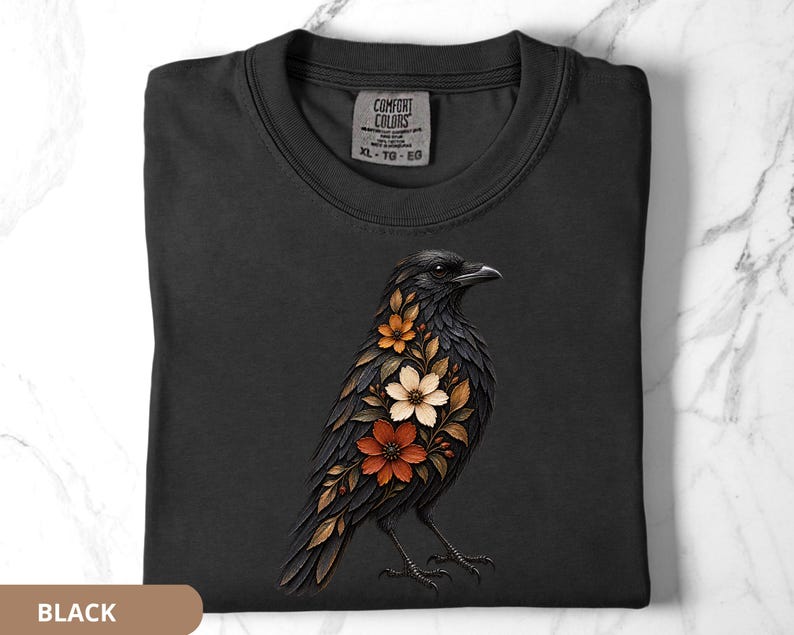 May include: A black t-shirt featuring a graphic of a crow with its chest filled with orange, white, and brown flowers and leaves. The shirt has a Comfort Colors brand label at the neckline with size XL-TG-EG printed on the interior fabric tag.