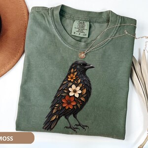 May include: A moss green crew neck t-shirt featuring a graphic of a black crow with orange, brown, and cream-colored flowers integrated into its plumage. The shirt has a Comfort Colors brand label at the inner neckline and short sleeves.