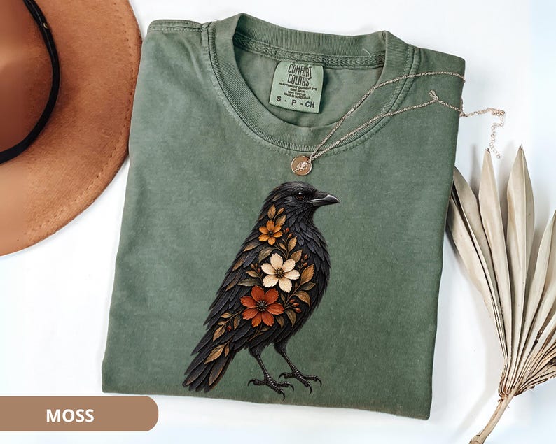 May include: A moss green crew neck t-shirt featuring a graphic of a black crow with orange, brown, and cream-colored flowers integrated into its plumage. The shirt has a Comfort Colors brand label at the inner neckline and short sleeves.