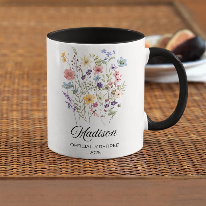 Op de afbeelding: Witte keramische mok met een zwarte binnenkant en handvat. De mok heeft een bloemenontwerp in pastelkleuren en de tekst "Madison Officially Retired 2025". De mok staat op een geweven oppervlak.