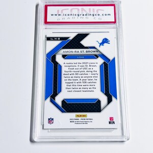 May include: A football trading card featuring Amon-Ra St. Brown of the Lions, encased in a protective holder. The card has blue and white accents with the Lions logo. Text includes player information and the Panini logo.