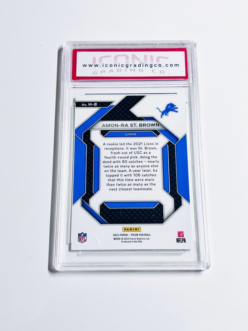 May include: A football trading card featuring Amon-Ra St. Brown of the Lions, encased in a protective holder. The card has blue and white accents with the Lions logo. Text includes player information and the Panini logo.