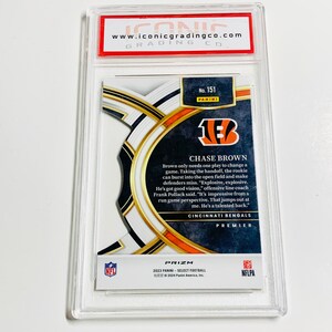 May include: A graded Chase Brown football card from the 2023 Panini Select Prizm series. The card features the Cincinnati Bengals logo and player information. Encased in a clear protective holder with the Iconic Grading Co. logo.