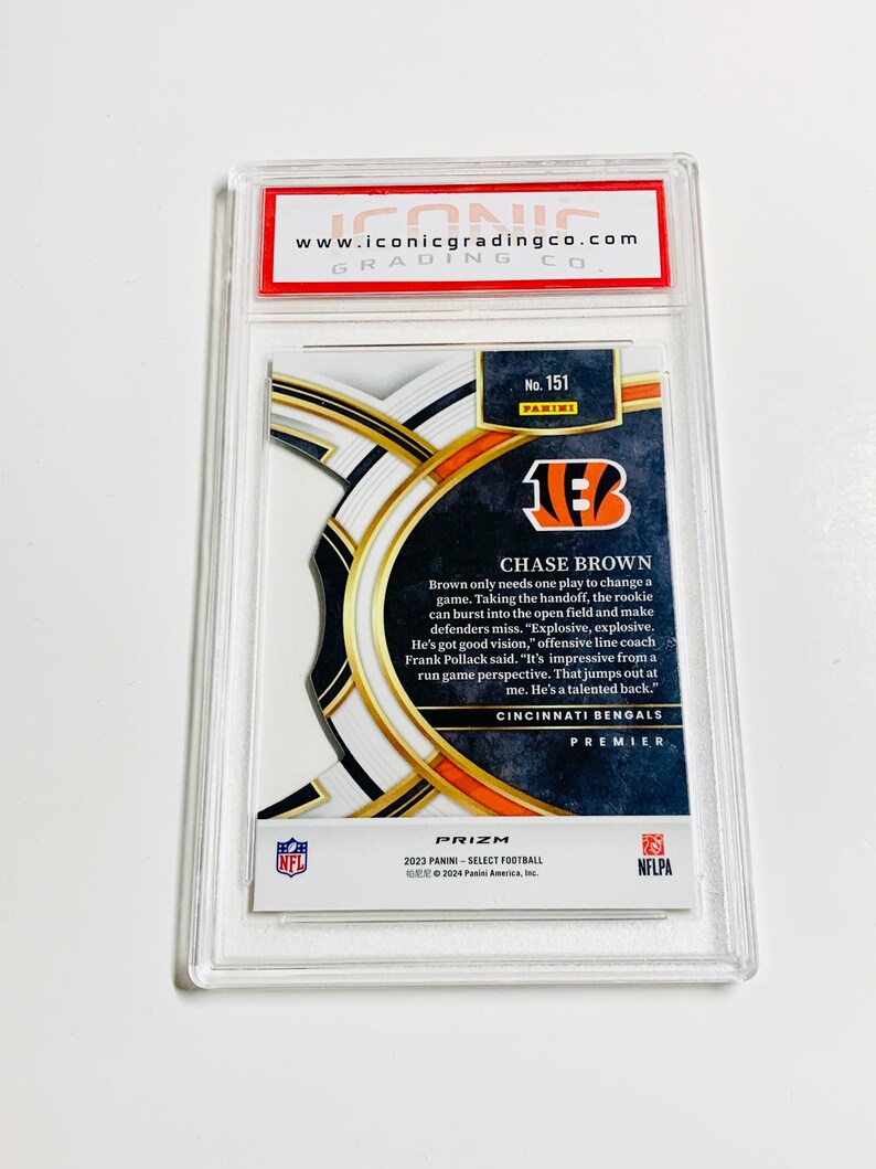 May include: A graded Chase Brown football card from the 2023 Panini Select Prizm series. The card features the Cincinnati Bengals logo and player information. Encased in a clear protective holder with the Iconic Grading Co. logo.