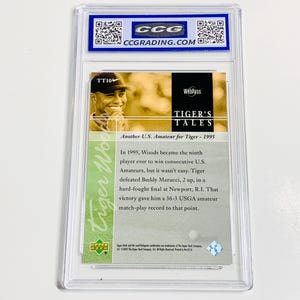 May include: A Tiger Woods trading card encased in a clear plastic holder. The card features a photo of Tiger Woods and text that reads "TIGER'S TALES" and "Another U.S. Amateur for Tiger - 1995". The card is graded by CCG.