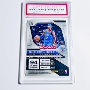K&ouml;nnte beinhalten: Eine Basketball-Sammelkarte mit Shai Gilgeous-Alexander in einem blauen Thunder-Trikot. Die Karte ist bewertet und in einem Schutzhalter eingeschlossen. Die Karte zeigt Spielerstatistiken, ein "Monopoly"-Logo und die Marke Panini.