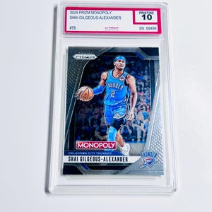 K&ouml;nnte beinhalten: Basketball-Sammelkarte mit Shai Gilgeous-Alexander im blauen Trikot der Oklahoma City Thunder. Die Karte ist mit "Pristine 10" bewertet und stammt aus der 2024 Prizm Monopoly-Serie. Die Karte ist in einer transparenten Schutzh&uuml;lle.