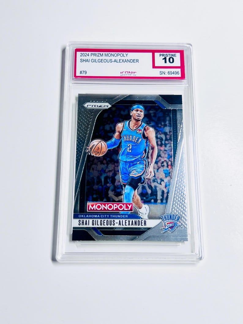 K&ouml;nnte beinhalten: Basketball-Sammelkarte mit Shai Gilgeous-Alexander im blauen Trikot der Oklahoma City Thunder. Die Karte ist mit "Pristine 10" bewertet und stammt aus der 2024 Prizm Monopoly-Serie. Die Karte ist in einer transparenten Schutzh&uuml;lle.