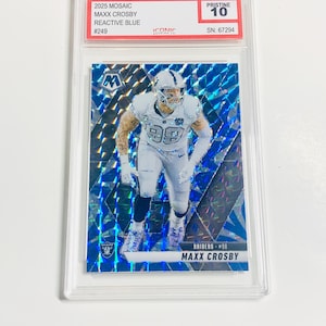 May include: A football trading card featuring Maxx Crosby in a white Raiders uniform. The card has a blue mosaic background and is graded Pristine 10. Text on the card includes "2025 Mosaic," "Reactive Blue," and "Raiders #98."