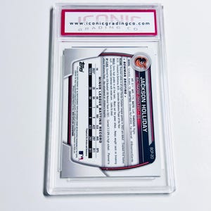 Puede incluir: Una tarjeta de b&eacute;isbol dentro de un estuche de pl&aacute;stico transparente. La tarjeta presenta a Jackson Holliday de los Orioles de Baltimore. La tarjeta tiene el logotipo de Topps y un registro de bateo. El soporte tiene una etiqueta roja y blanca con el sitio web "www.iconicgradingco.com".