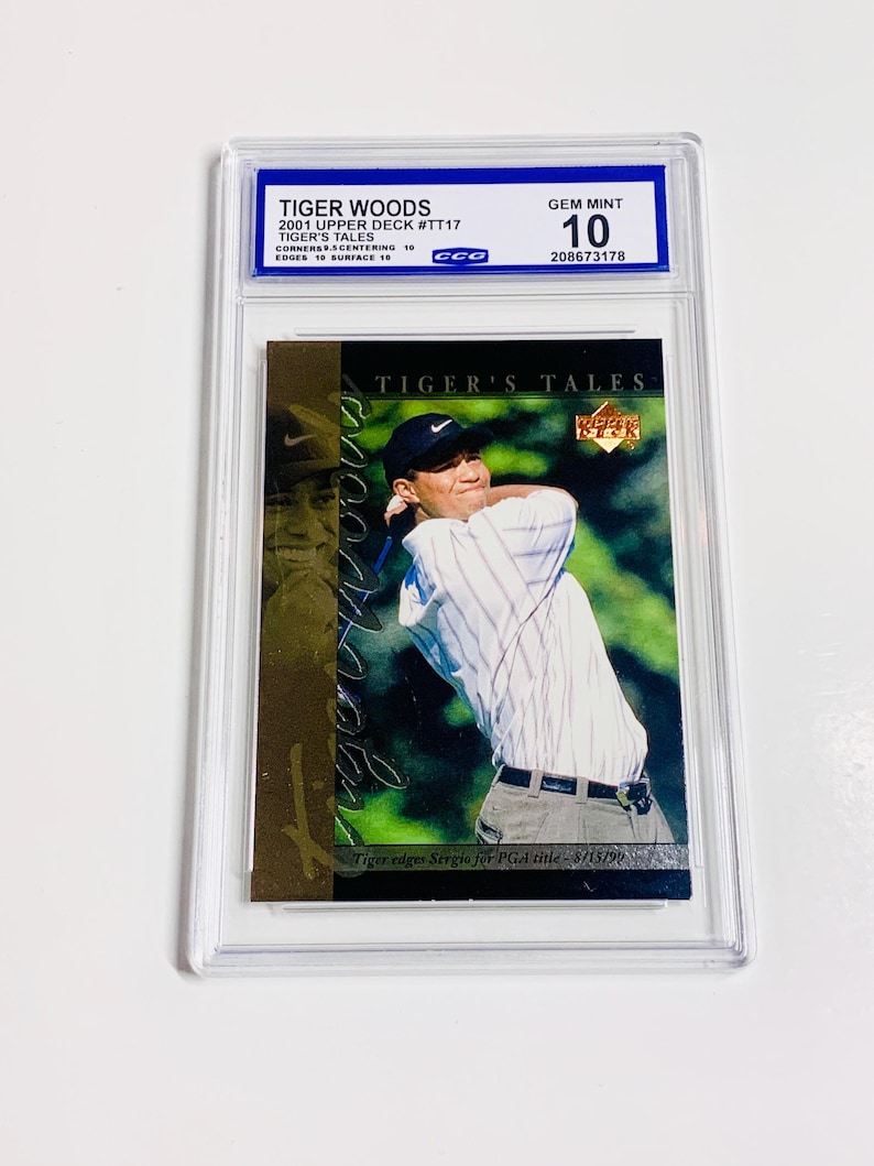 May include: A Tiger Woods 2001 Upper Deck #TT17 trading card, graded Gem Mint 10. The card features a photo of Tiger Woods in action, with the text "TIGER'S TALES" and "Tiger edges Sergio for PGA title - 8/15/99". The card is encased in a protective holder.