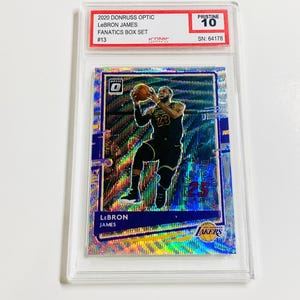 May include: A basketball trading card featuring LeBron James in a Los Angeles Lakers uniform, holding a basketball. The card is encased in a protective holder and labeled "2020 Donruss Optic" and "Pristine 10". The card has a holographic design.
