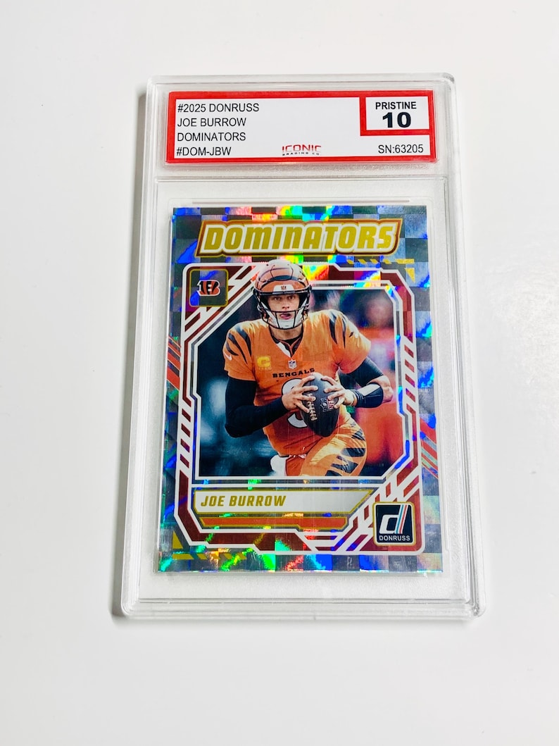 Może przedstawiać: Karta do gry w futbol amerykański Joe Burrow z serii Donruss Dominators, oceniona na Pristine 10. Karta przedstawia zdjęcie Burrowa w jego mundurze Bengals, trzymającego piłkę do futbolu. Karta jest zamknięta w ochronnym uchwycie.