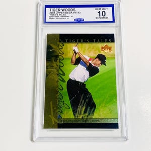 Op de afbeelding: Een Tiger Woods 2001 Upper Deck #TT13 trading card, beoordeeld als GEM MINT 10, in een beschermende houder. De kaart toont Tiger Woods die golf speelt en de tekst "TIGER'S TALES".