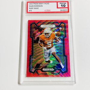 Puede incluir: Una tarjeta de fútbol coleccionable roja e iridiscente con Bijan Robinson con el uniforme de los Texas Longhorns. La tarjeta está calificada como Pristine 10 e incluye el texto "2024 Prizm Draft Picks" y "Ruby Wave".