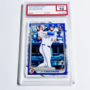 May include: A baseball trading card of Jac Caglianone in a protective case. The card features a player in a Kansas City Royals uniform. The card is graded "Pristine 10" and includes the text "2024 Bowman Draft".