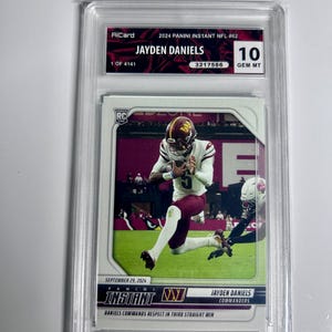 Puede incluir: Una tarjeta comercial de 2024 Panini Instant NFL #62 calificada con Jayden Daniels de los Commanders. La tarjeta est&aacute; calificada como 10 GEM MT y est&aacute; encerrada en un soporte protector. La tarjeta muestra a Daniels en acci&oacute;n, con el texto "DANIELS COMMANDS RESPECT IN THIRD STRAIGHT WIN".