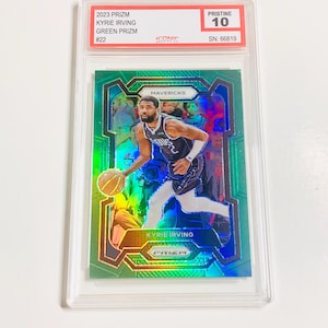 May include: A 2023 Prizm Kyrie Irving Green Prizm basketball card, graded Pristine 10, encased in a clear protective holder. The card features a vibrant image of Kyrie Irving in action, with the Mavericks logo visible.