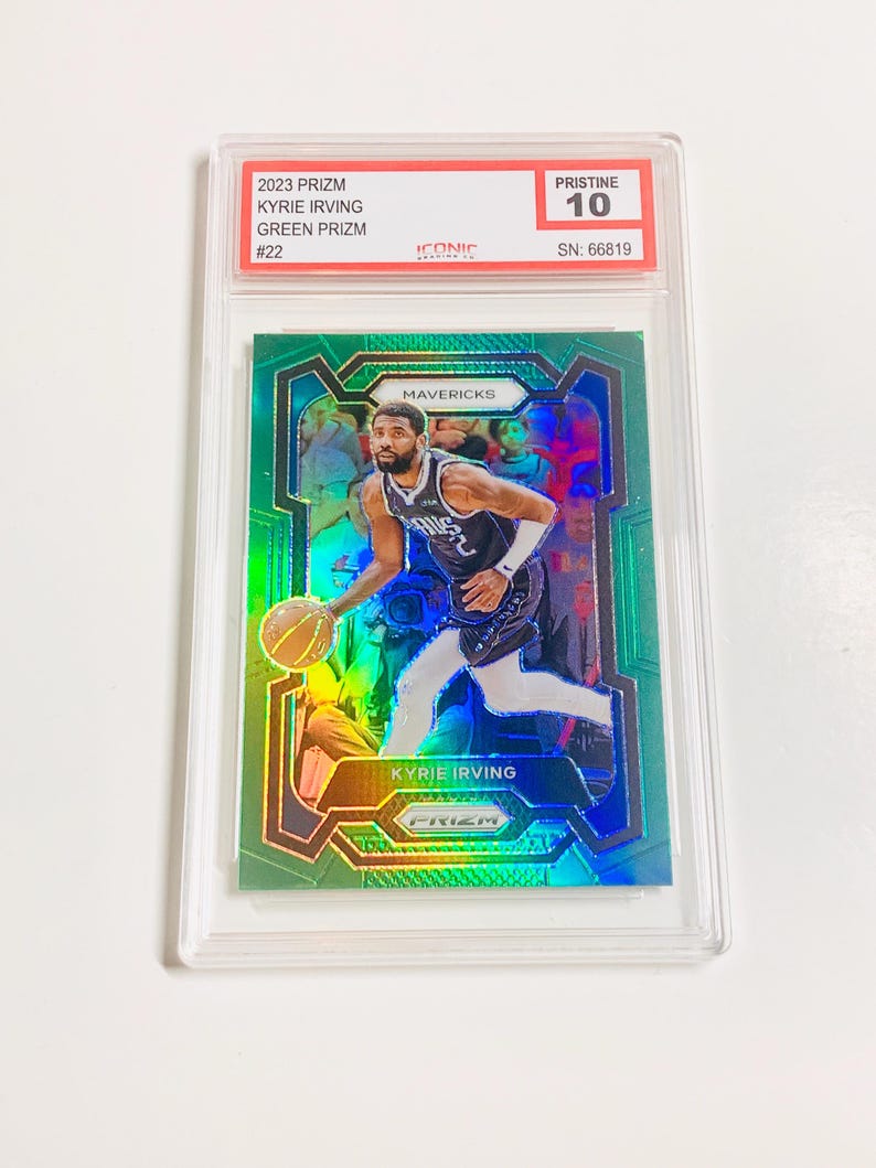 May include: A 2023 Prizm Kyrie Irving Green Prizm basketball card, graded Pristine 10, encased in a clear protective holder. The card features a vibrant image of Kyrie Irving in action, with the Mavericks logo visible.