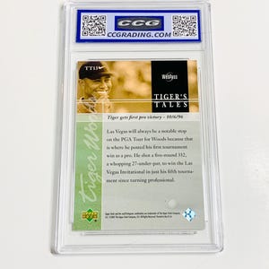 Op de afbeelding: Een Tiger Woods-verzamelkaart in een doorzichtige plastic houder. De kaart toont een foto van Woods en de tekst "Tiger's Tales" en "Tiger gets first pro victory - 10/6/96". De kaart is beoordeeld door CCG.