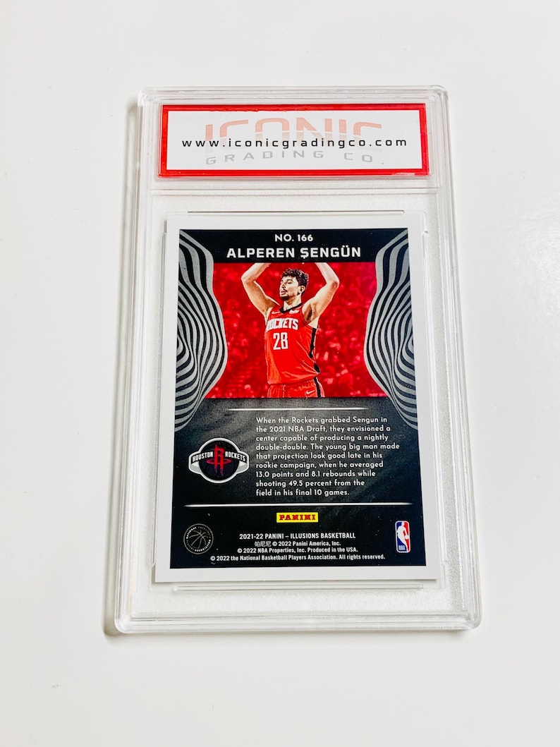 Puede incluir: Una tarjeta de baloncesto de Alperen Sengun calificada de la serie Panini Illusions 2021-22. La tarjeta presenta una foto de Sengun con una camiseta roja de los Rockets. La tarjeta est&aacute; dentro de un soporte de pl&aacute;stico transparente con el logotipo de Iconic Grading Co.