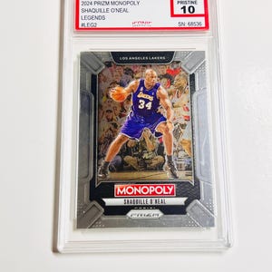 May include: A basketball trading card featuring Shaquille O'Neal in a Los Angeles Lakers uniform. The card is from the 2024 Prizm Monopoly Legends series and is graded Pristine 10. The card is encased in a clear protective holder.
