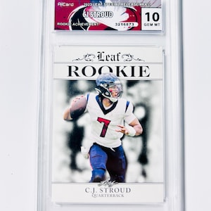 Puede incluir: Una tarjeta coleccionable deportiva calificada con C.J. Stroud, un mariscal de campo, con uniforme blanco y azul. La tarjeta est&aacute; etiquetada como "Rookie" e incluye el texto "2023 Leaf Special Release". La tarjeta est&aacute; calificada con un 10, GEM MT. La tarjeta est&aacute; dentro de un soporte de pl&aacute;stico transparente.