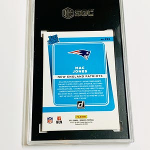 May include: A graded Mac Jones football card from the 2021 Panini Donruss series. The card features a blue and white design with the New England Patriots logo and player information. The card is encased in a black holder with the SGC logo.
