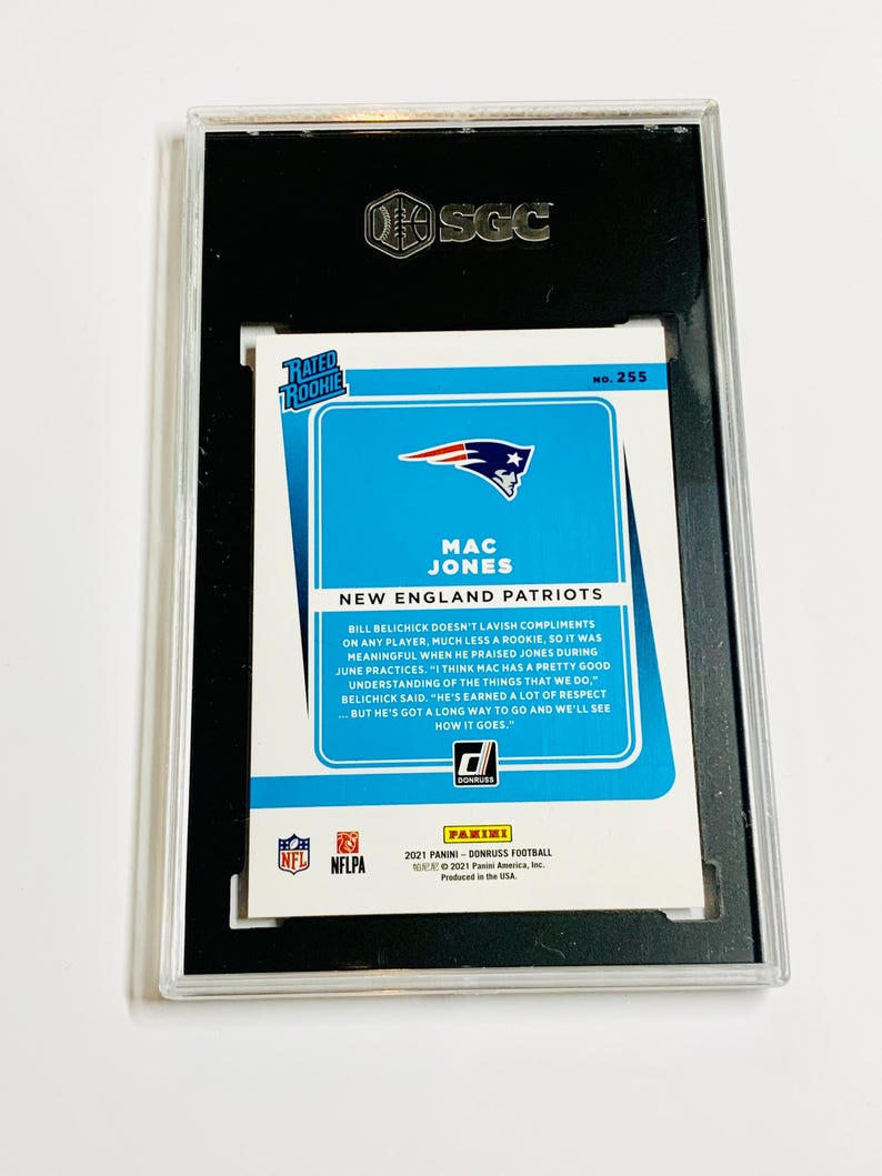 May include: A graded Mac Jones football card from the 2021 Panini Donruss series. The card features a blue and white design with the New England Patriots logo and player information. The card is encased in a black holder with the SGC logo.