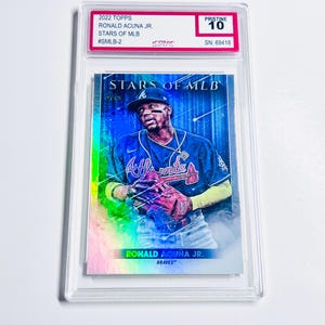 May include: A baseball card featuring Ronald Acu&ntilde;a Jr. of the Atlanta Braves, from the 2022 Topps Stars of MLB series. The card is graded Pristine 10 and encased in a clear protective holder. The card has a holographic design with the player in action.