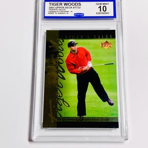 Puede incluir: Una tarjeta coleccionable de Tiger Woods de la serie Upper Deck de 2001, calificada como Gem Mint 10. La tarjeta presenta a Woods con una camisa roja y pantalones negros, en pleno swing en un campo de golf. La tarjeta está en un soporte protector.