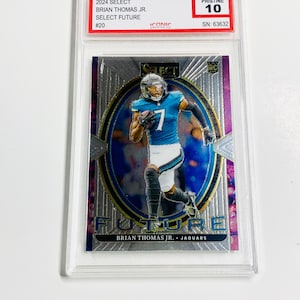 May include: A trading card featuring Brian Thomas Jr. in a teal and white uniform, numbered 7. The card is encased in a clear protective holder. The card's text includes "2024 Select", "Select Future", and "Jaguars". The card is graded "Pristine 10".