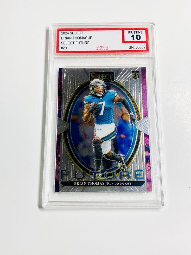 May include: A trading card featuring Brian Thomas Jr. in a teal and white uniform, numbered 7. The card is encased in a clear protective holder. The card's text includes "2024 Select", "Select Future", and "Jaguars". The card is graded "Pristine 10".