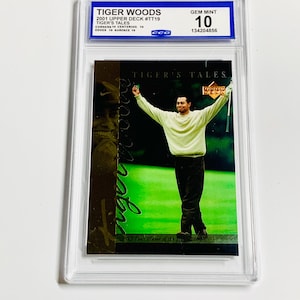 Puede incluir: Tarjeta coleccionable de Tiger Woods de 2001 Upper Deck, con Woods en un campo de golf verde con los brazos levantados. La tarjeta está en un soporte de plástico transparente, calificada GEM MINT 10. De la serie 'Tiger's Tales'.