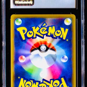 Pu&ograve; includere: Una carta collezionabile Pok&eacute;mon valutata, racchiusa in un supporto di plastica trasparente. Il retro della carta presenta il logo Pok&eacute;mon in giallo e la parola "Pok&eacute;mon" in giallo. La carta ha uno sfondo blu con globi colorati.
