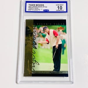 Op de afbeelding: Een beoordeelde ruilkaart met Tiger Woods uit de 2001 Upper Deck set. De kaart toont Woods op een golfbaan, met de tekst "TIGER'S TALES" en "A memorable Memorial for Tiger - 5/28/00". De kaart is beoordeeld met GEM MINT 10.