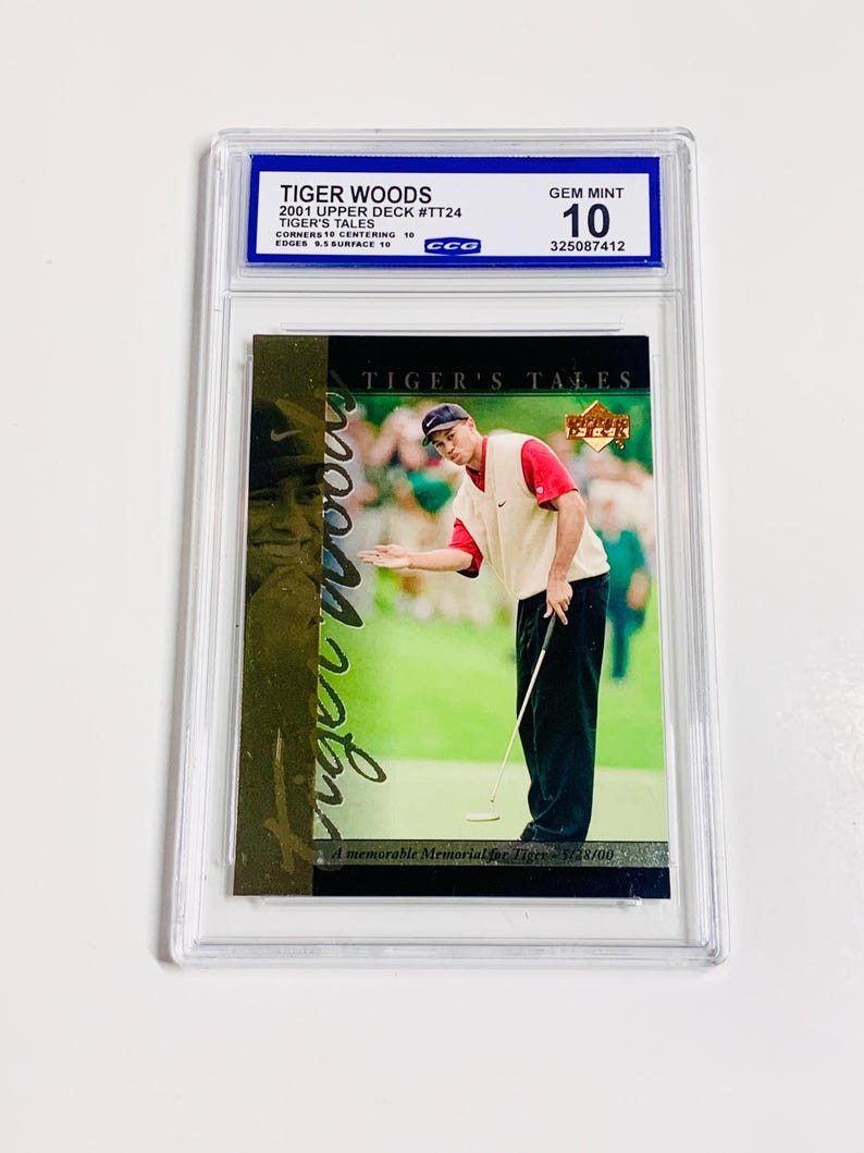 Op de afbeelding: Een beoordeelde ruilkaart met Tiger Woods uit de 2001 Upper Deck set. De kaart toont Woods op een golfbaan, met de tekst "TIGER'S TALES" en "A memorable Memorial for Tiger - 5/28/00". De kaart is beoordeeld met GEM MINT 10.