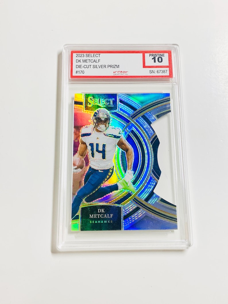 May include: A 2023 Select DK Metcalf Die-Cut Silver Prizm trading card, graded Pristine 10. The card features a player in a Seahawks uniform, with the number 14. The card is encased in a clear protective holder.