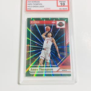 Op de afbeelding: Een basketbal trading card met Amen Thompson in actie, met een groen holografisch ontwerp. De kaart is beoordeeld met Pristine 10 en zit in een beschermende houder. De kaart leest "2024 Donruss" en "Holo Green Laser".