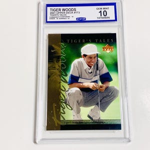 Puede incluir: Una tarjeta coleccionable de Tiger Woods del set Upper Deck 2001, calificada como Gem Mint 10. La tarjeta presenta a Tiger Woods en una pose de golf, con el texto "TIGER'S TALES". La tarjeta está dentro de un soporte protector.
