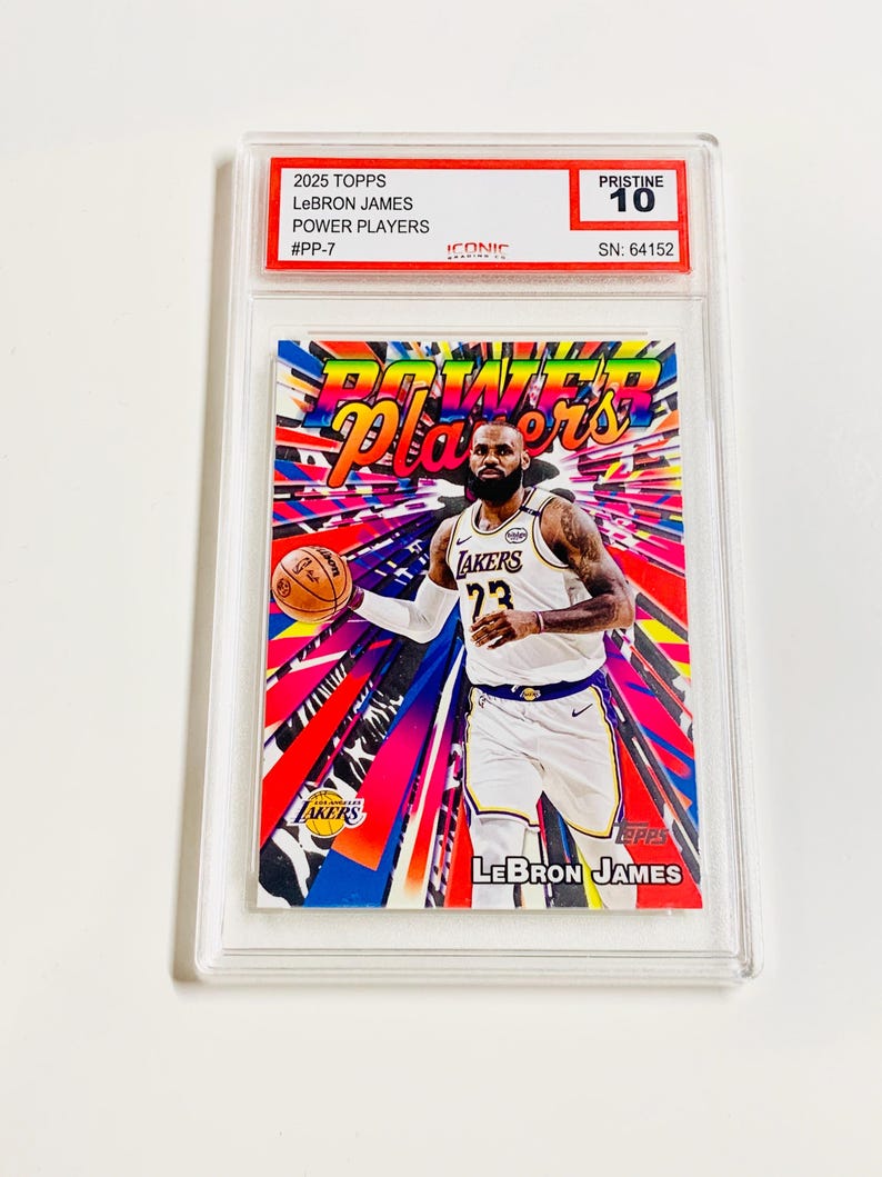 K&ouml;nnte beinhalten: Eine 2025 Topps LeBron James Power Players Sammelkarte, bewertet mit Pristine 10, in einer Schutzh&uuml;lle. Die Karte zeigt LeBron James im Lakers-Trikot vor einem leuchtenden, farbenfrohen Hintergrund. Der Text auf der Karte enth&auml;lt "Power Players" und "LeBron James".