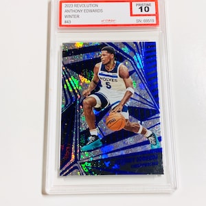Op de afbeelding: Een basketbal trading card met Anthony Edwards in een Minnesota Timberwolves uniform, dribbelend met een basketbal. De kaart zit in een beschermende houder en heeft een blauw en zilver holografisch ontwerp. De kaart is gelabeld "2023 Revolution" en "Pristine 10".