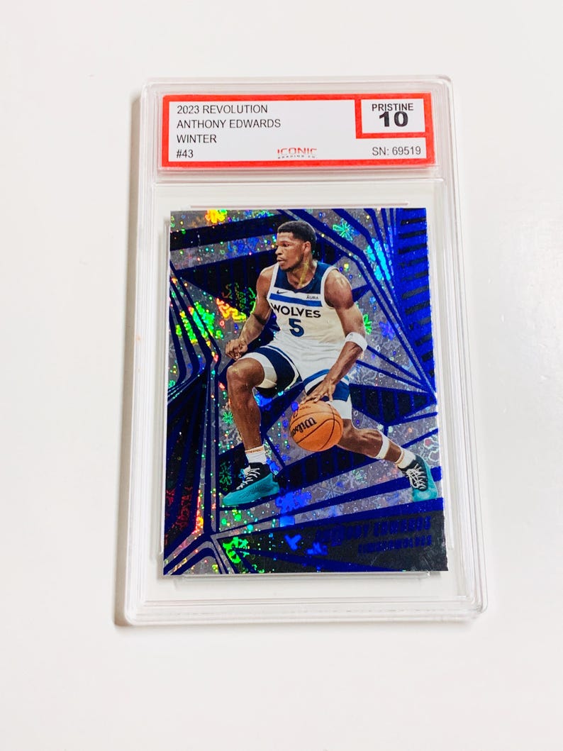 Op de afbeelding: Een basketbal trading card met Anthony Edwards in een Minnesota Timberwolves uniform, dribbelend met een basketbal. De kaart zit in een beschermende houder en heeft een blauw en zilver holografisch ontwerp. De kaart is gelabeld "2023 Revolution" en "Pristine 10".